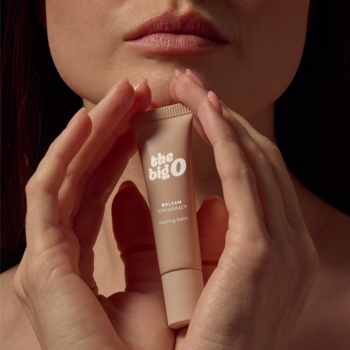 Zbliżenie na beżowy balsam the big O cooling balm do seksu oralnego, delikatnie trzymany w kobiecych dłoniach, usta w tle.