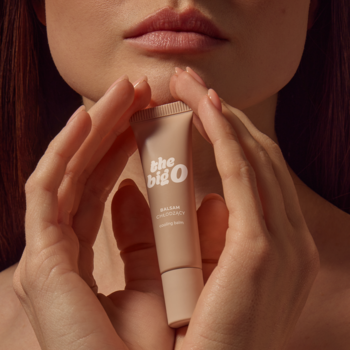 Beżowy balsam chłodzący The Big O w matowej tubce, trzymany w kobiecych dłoniach, na tle ust. Intimacy care.