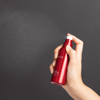 Czerwony spray do twarzy Suntie, rozpylający delikatną mgiełkę, trzymany w dłoni na ciemnym tle.