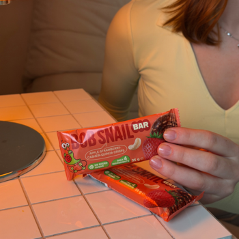 Zbliżenie na baton owocowy Bob Snail Bar jabłkowo-truskawkowy w czerwonym opakowaniu, trzymany w dłoniach nad stołem.