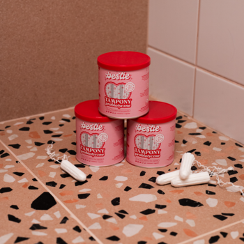 Trzy różowe pojemniki Bestie Tampony Probiotyczne Medium w stosie na nakrapianej posadzce, obok leżą białe tampony.