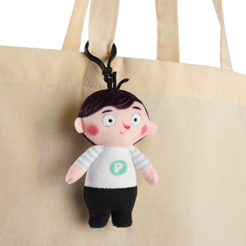 Pluszowy breloczek Pucio w białym T-shircie z zielonym logo 'P', z czarnymi włosami, zawieszony na beżowej torbie.