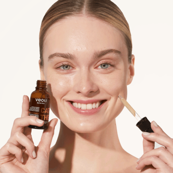Kobieta z uśmiechem aplikuje kropelki brązujące Veoli Botanica Catch The Sun na policzek, trzymając butelkę.