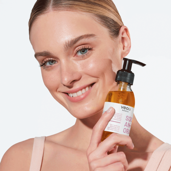 Uśmiechnięta kobieta trzyma olejek do demakijażu Veoli Botanica Squeeze An Orange w przezroczystej butelce z pompką.