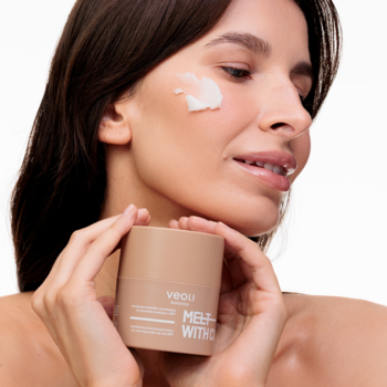 Kobieta z kremem na policzku trzyma beżowe masełko Veoli Botanica Melt With Comfort, nawilżające do demakijażu.