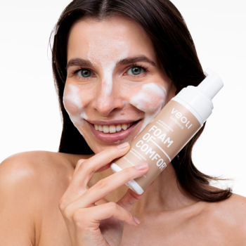 Uśmiechnięta kobieta z białą pianką na twarzy, trzymająca beżową butelkę pianki Veoli Botanica Foam Of Comfort.