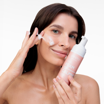 Kobieta aplikująca kojącą piankę Veoli Botanica Foam Of Calmness na twarz, trzymająca różową butelkę.