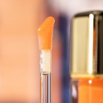 Zbliżenie na aplikator pomarańczowego błyszczyka do ust z miękką, włochatą końcówką, na rozmytym tle butelki.