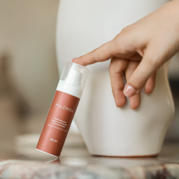 Brązowa butelka serum rozświetlającego POLE MULA z witaminą C, którą dotyka dłoń, przy białej wazie.