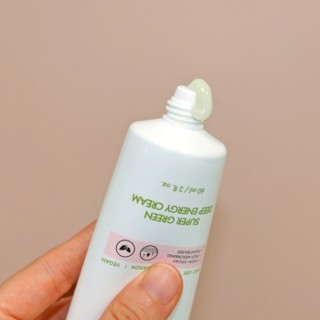 Zbliżenie na dłoń trzymającą białą tubkę SUPER GREEN DEEP ENERGY CREAM, z której wyciskany jest gęsty, jasnozielony krem.