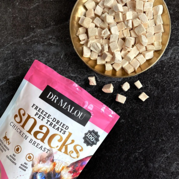 Dr Malou Liofilizowane przysmaki z kurczaka dla kota, różowa torebka i złota miseczka z kostkami na ciemnym tle.