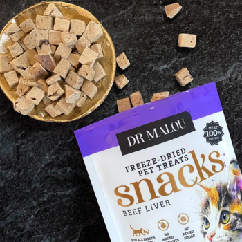 Opakowanie Dr Malou Freeze-Dried Beef Liver snacks dla kota, obok miseczka i rozsypane kostki przysmaków na ciemnym blacie.