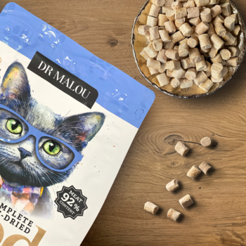 Opakowanie karmy Dr Malou liofilizowanej (kaczka/jagnięcina) z kotem w okularach, obok granulki na drewnie.