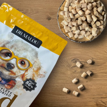 Dr Malou Kurczak z dynią, liofilizowana karma dla psa. Biało-żółte opakowanie i rozsypane granulki na drewnie.