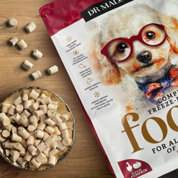 Biało-czerwona torba karmy liofilizowanej Dr Malou Kaczka z dynią, z psem w okularach, obok sucha karma w misce.