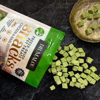 Otwarte opakowanie Dr Malou z liofilizowanymi przysmakami dla kota (kurczak, trawa jęczmienna), zielone kostki na czarnym blacie.