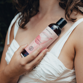Żel do mycia ciała Organique Naturals Sensitive magnolia, w ciemnej butelce z różową etykietą, trzymany przy klatce piersiowej.