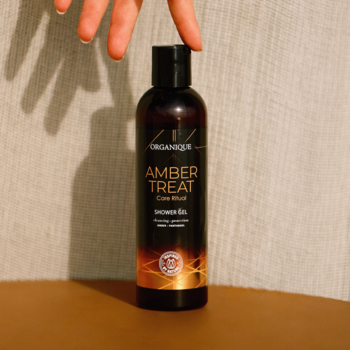 ORGANIQUE Amber Treat Shower Gel w ciemnej butelce z pomarańczową etykietą, trzymany przez dłoń. Nawilża, ujędrnia.