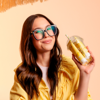 Uśmiechnięta kobieta w żółtym stroju trzyma tropikalny zestaw Fluff Coco-Bahama Smoothie w transparentnym kubku.