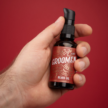 Męska dłoń trzyma ciemną butelkę olejku do brody GroomeN z pompką, czerwoną etykietą i złotym detalem, na czerwonym tle.