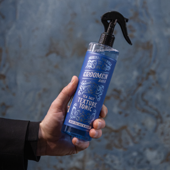 Groomen Sea Salt Texture Tonic AQUA, niebieski spray do włosów, butelka z atomizerem trzymana w dłoni.