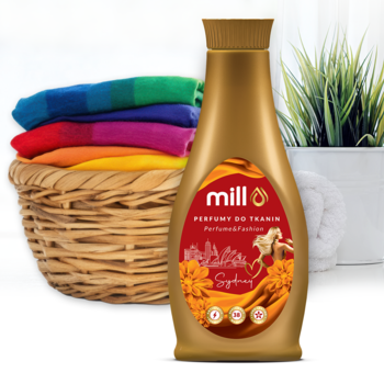 MILL Perfumy do tkanin SYDNEY 750 ml, złota butelka z czerwoną etykietą, w tle kosz barwnych ręczników.