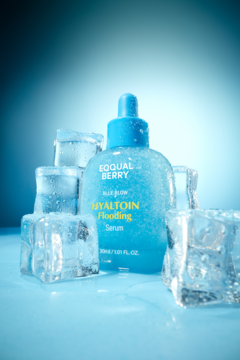EQQUAL BERRY HYALTOIN FLOODING Serum, niebieska butelka z kroplomierzem, otoczona lśniącymi kostkami lodu.