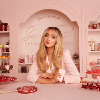 Sabrina Carpenter w różowym żakiecie promuje perfumy Sweet Tooth, uśmiechając się przy ladzie pełnej słodyczy.