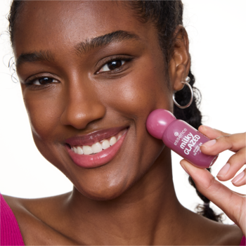 Uśmiechnięta kobieta trzyma różowy, zaokrąglony balsam essence milky GLAZED 04 Berry Au Lait przy policzku.