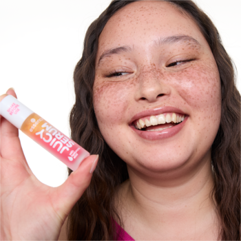 Uśmiechnięta kobieta z piegami prezentuje błyszczyk do ust Juicy Serum w przezroczystej tubce z różową etykietą.