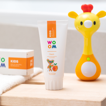 Biała tubka pasty WOOM KIDS PEACH z pomarańczową etykietą i brzoskwinią, obok żółta zabawka żyrafa.