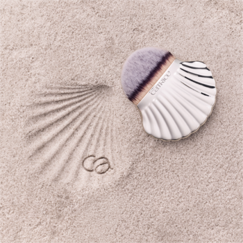 Pędzel Catrice Seashell do twarzy i ciała, biała muszla z szarym włosiem, na piasku z odciskiem i pierścionkami.