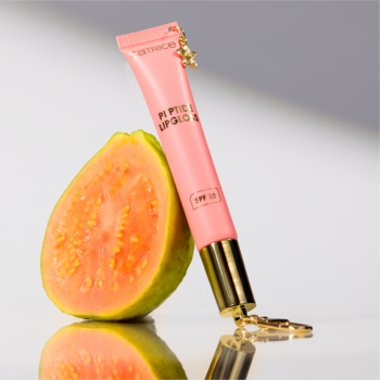 Różowy błyszczyk Catrice Guava Jam SPF 25 z ozdobną zakrętką, stojący obok połówki guawy na refleksyjnym tle.