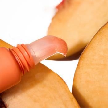 Aplikator błyszczyka Catrice Peach Iced Tea, o brzoskwiniowym odcieniu i mokrym wykończeniu, obok plastrów brzoskwini.