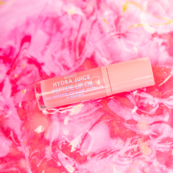 Hydra Juice Glossing Lip Oil, brzoskwiniowy olejek do ust w tubce, leży na jaskrawym różowo-białym, wirującym tle.