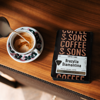 Opakowanie kawy ziarnistej Coffee & Sons Brazylia Diamantina na drewnie, obok filiżanka z wlewanym mlekiem.