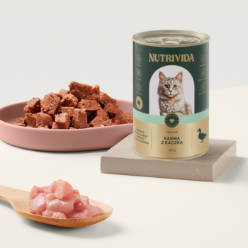 Puszka karmy Nutrivida dla kota z kaczką na podwyższeniu, obok miska pasztetu i surowe kawałki mięsa na łyżce.