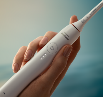 Biała elektryczna szczoteczka Philips Sonicare, widoczny przycisk zasilania i diody LED, trzymana w dłoni na jasnym tle.