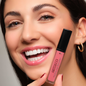 Uśmiechnięta kobieta z błyszczącymi ustami trzyma nawilżający błyszczyk Artdeco Hydra Lip Booster, Translucent Mountain Rose.