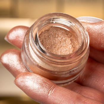 Glamtes Miami Beach pigment w otwartym, szklanym słoiczku, beżowo-szampański, ze złocistym blaskiem, trzymany w dłoni.
