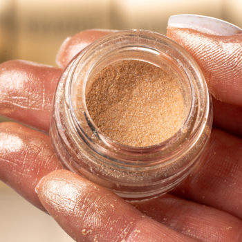 Złoty sypki pigment GLAMTES Liquid Gold 04 w otwartym szklanym słoiczku, trzymany w dłoni. Rozświetlający.