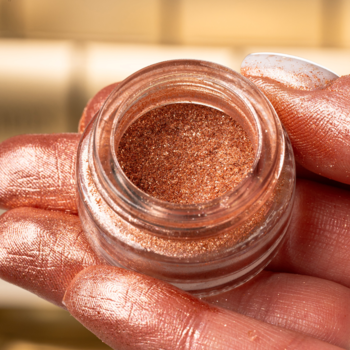 Makijażowy pigment Glamtes Rose Gold w szklanym słoiczku, trzymany w dłoni, z iskrzącym różowo-złotym połyskiem.