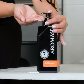Dłonie dozujące Kojący szampon AROMASE Dry Scalp z czarnej butelki z pompką, widoczna liczba 7 i pomarańczowa etykieta.