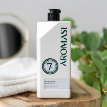 Biała, kwadratowa butelka ziołowego szamponu Aromase Herbal Daily Essential, z zielonym napisem, na drewnie.