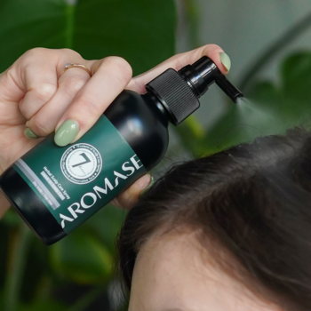 Dłoń w zielonym lakierze spryskuje AROMASE Ziołową wcierką nawilżającą ciemne włosy przy skórze głowy.
