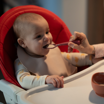 Uśmiechnięte dziecko w czerwonym krzesełku jest karmione łyżeczką z małej miseczki przez dorosłego.