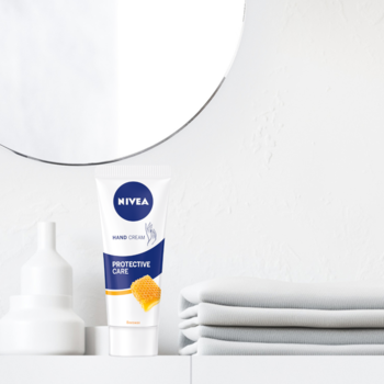 NIVEA Protective Care Krem do rąk, biała tubka z miodem, stoi na białej półce w łazience z lustrem i ręcznikami.