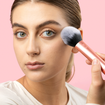 Kobieta nakłada puder puszystym pędzlem Real Techniques Powder Brush z miedzianą rączką, na różowym tle.