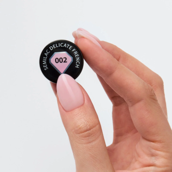Kobieca dłoń z delikatnymi, różowymi paznokciami trzyma czarny wzornik Semilac 002 Delicate French, ukazujący mleczny, blady róż.
