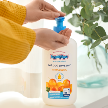 Ręce osoby dozującej z białej butelki Bambino Rodzina żel pod prysznic Mirabelka, 1000 ml, na jasnym tle.